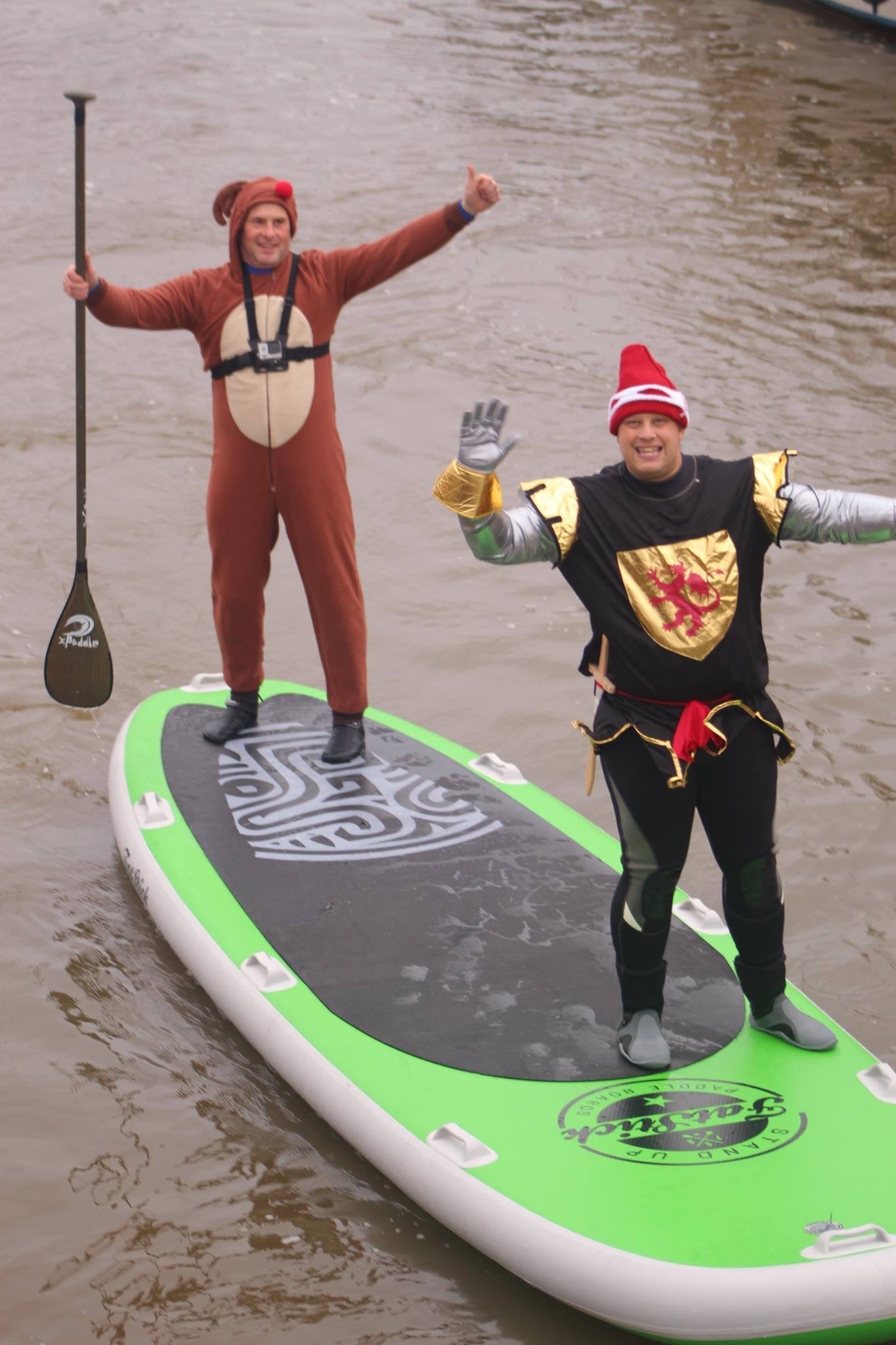 Two men in costumes, one dressed as a bear and one as a knight, standing on a green and black standing paddle board in the water.