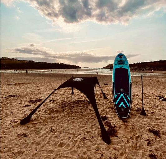 Black Fatstick Beach Tent and blue paddleboard on a beach at sunset with ocean and hills.