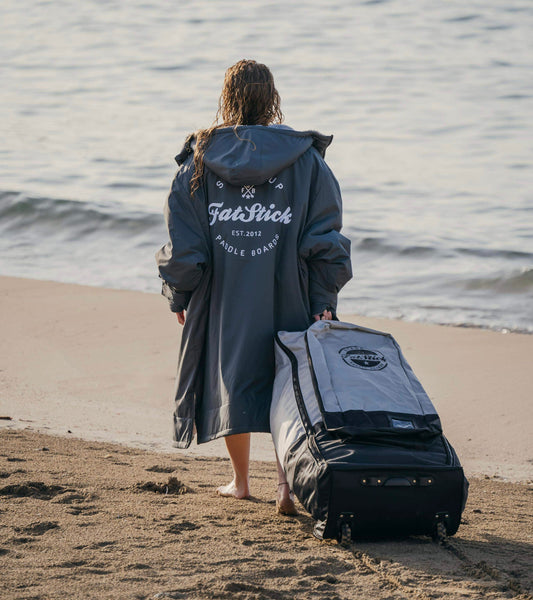 Woman in grey robe pulling a FatStick Wheeled Storage Bag Backpack along a beach towards the ocean.