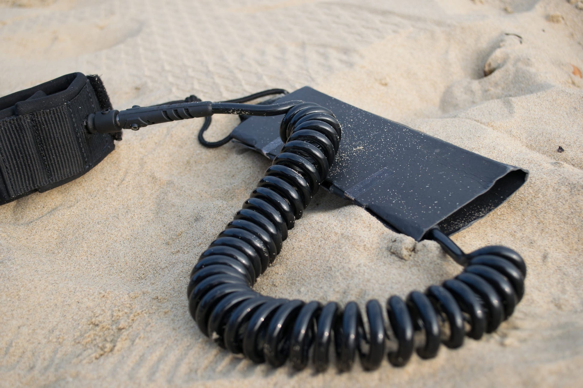 FatStick Boards black coiled 10ft SUP ankle leash lying on a sandy beach