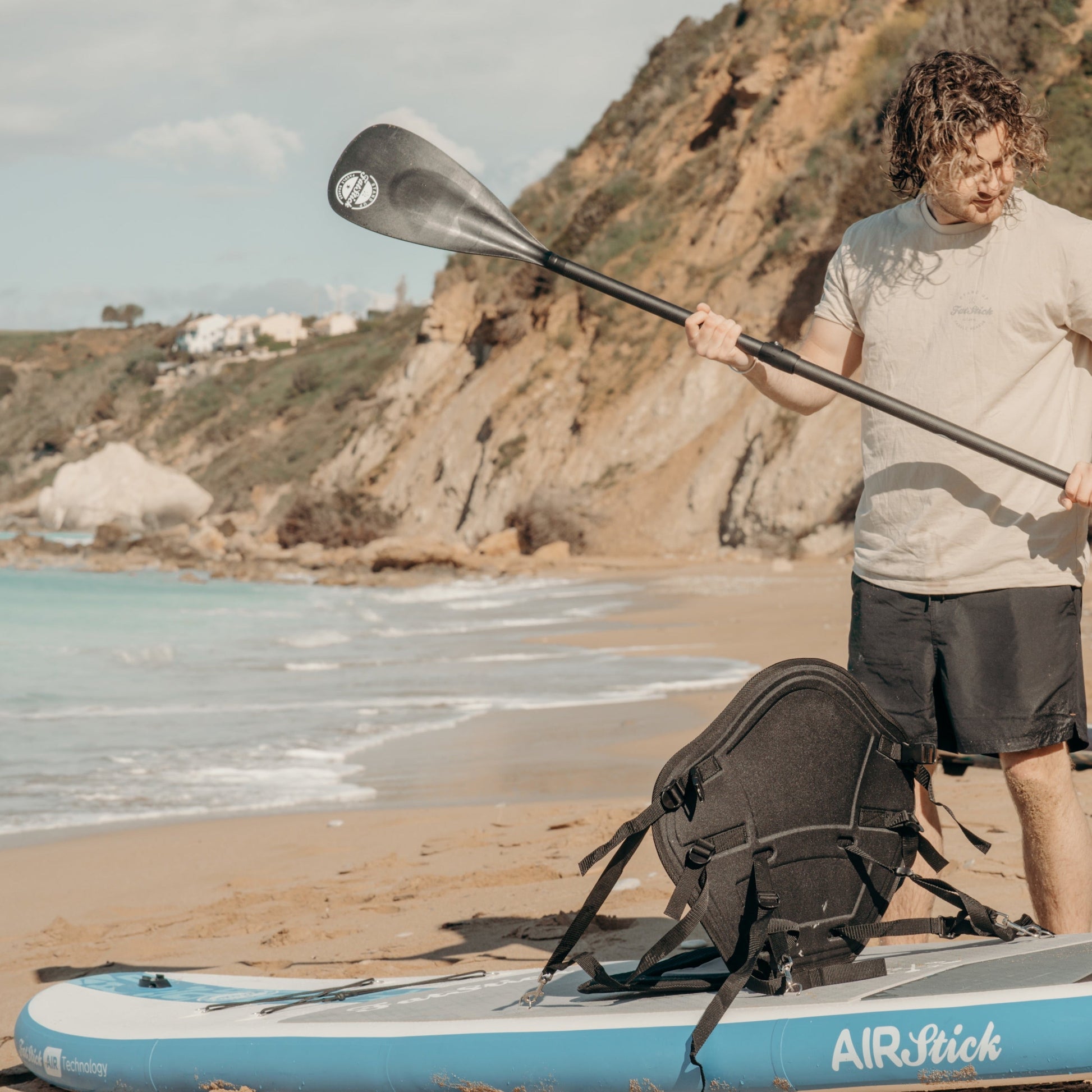 Man holding a paddle on a beach next to the AirStick inflatable paddle board with kayak seat attachment.
