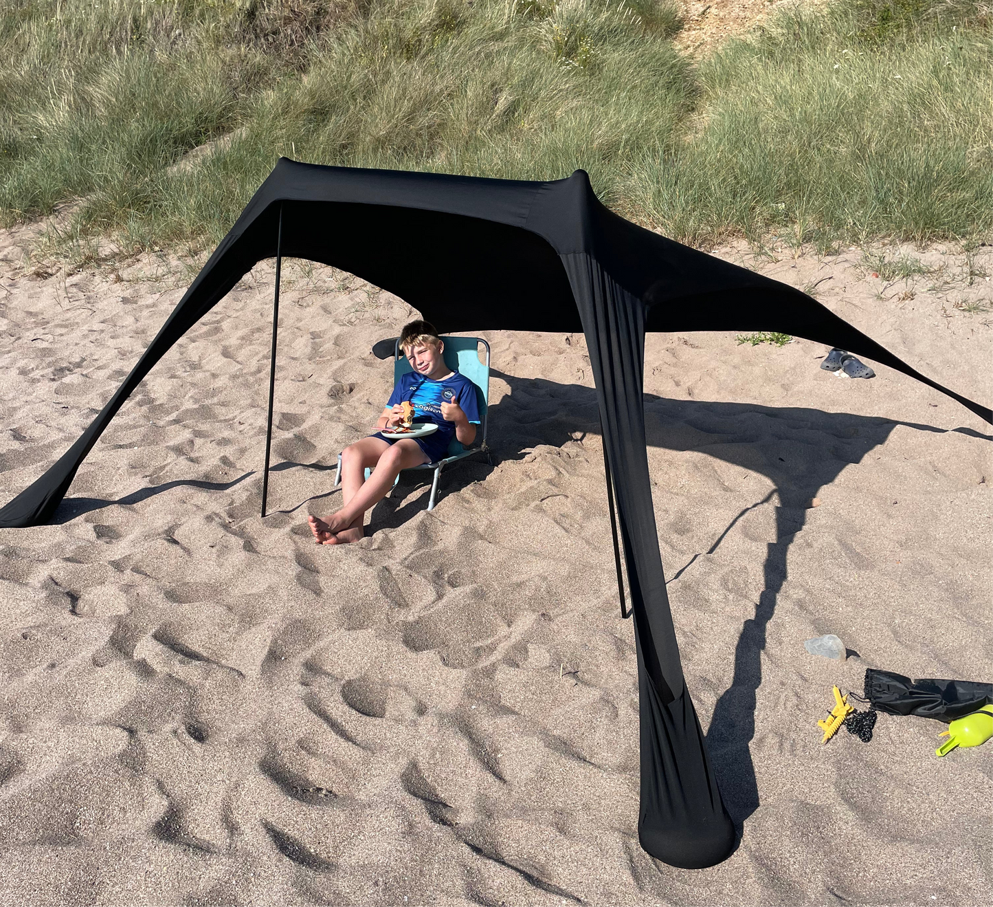 Child eating a snack under the black Fatstick Beach Tent sun shade on a sandy beach.