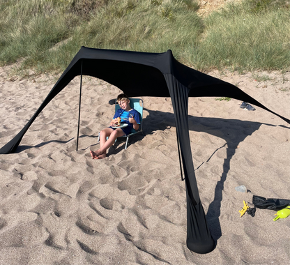 Child eating a snack under the black Fatstick Beach Tent sun shade on a sandy beach.