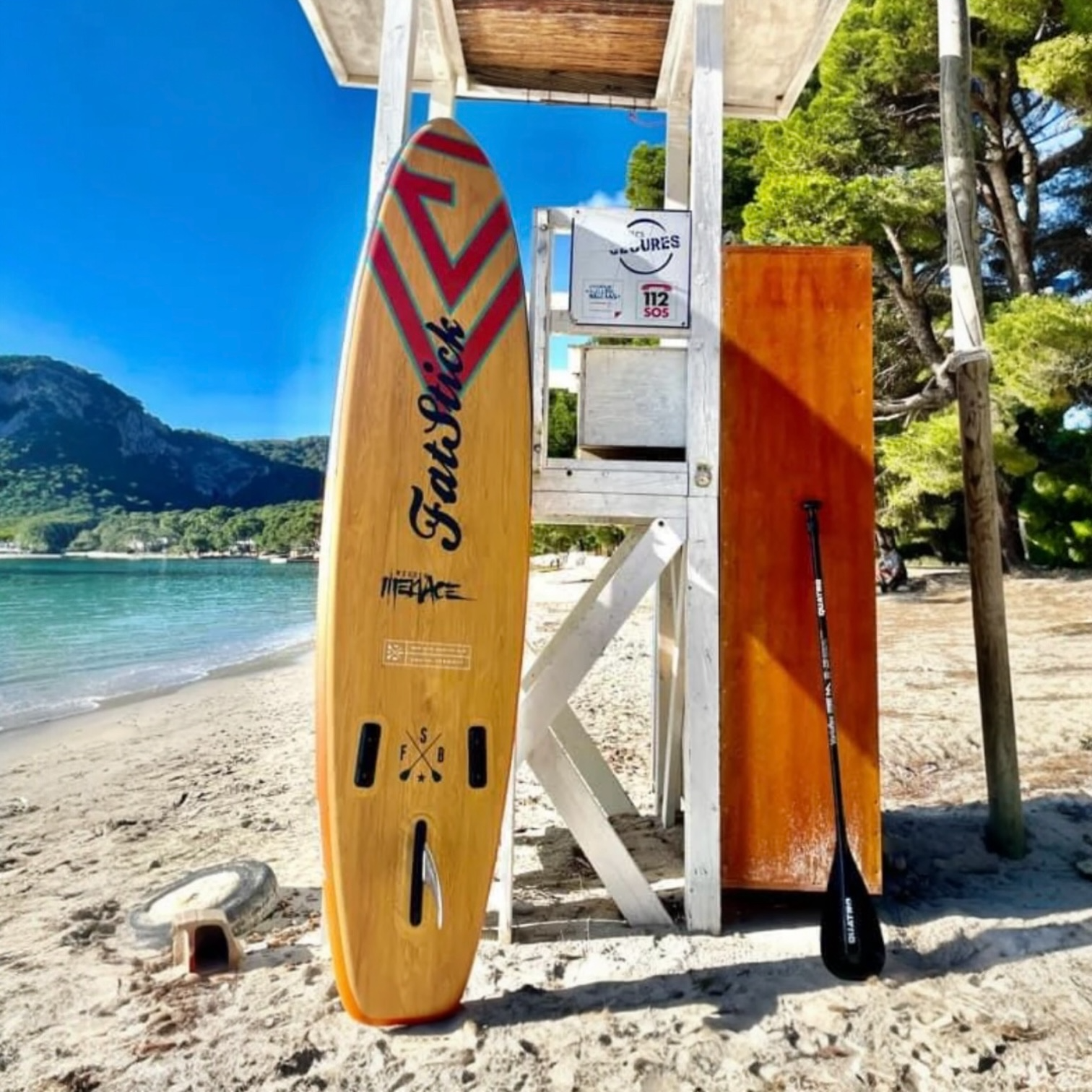 FatStick Wooden Menace 11'2 inflatable paddle board leaning against a white lifeguard stand on a sandy beach with blue skies.