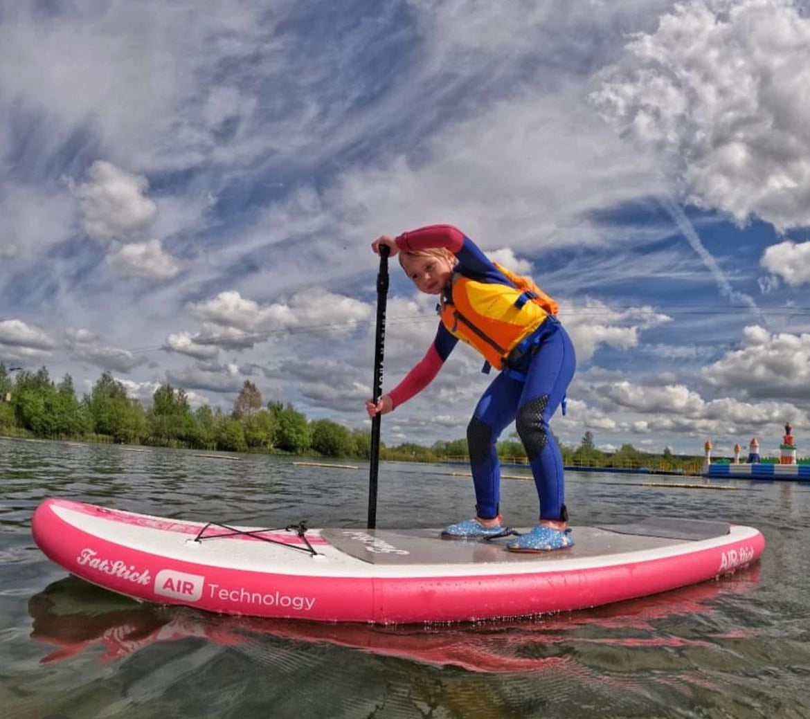 Smiling girl in a life vest on a pink Fatstick Gromstick inflatable paddle board on the water.