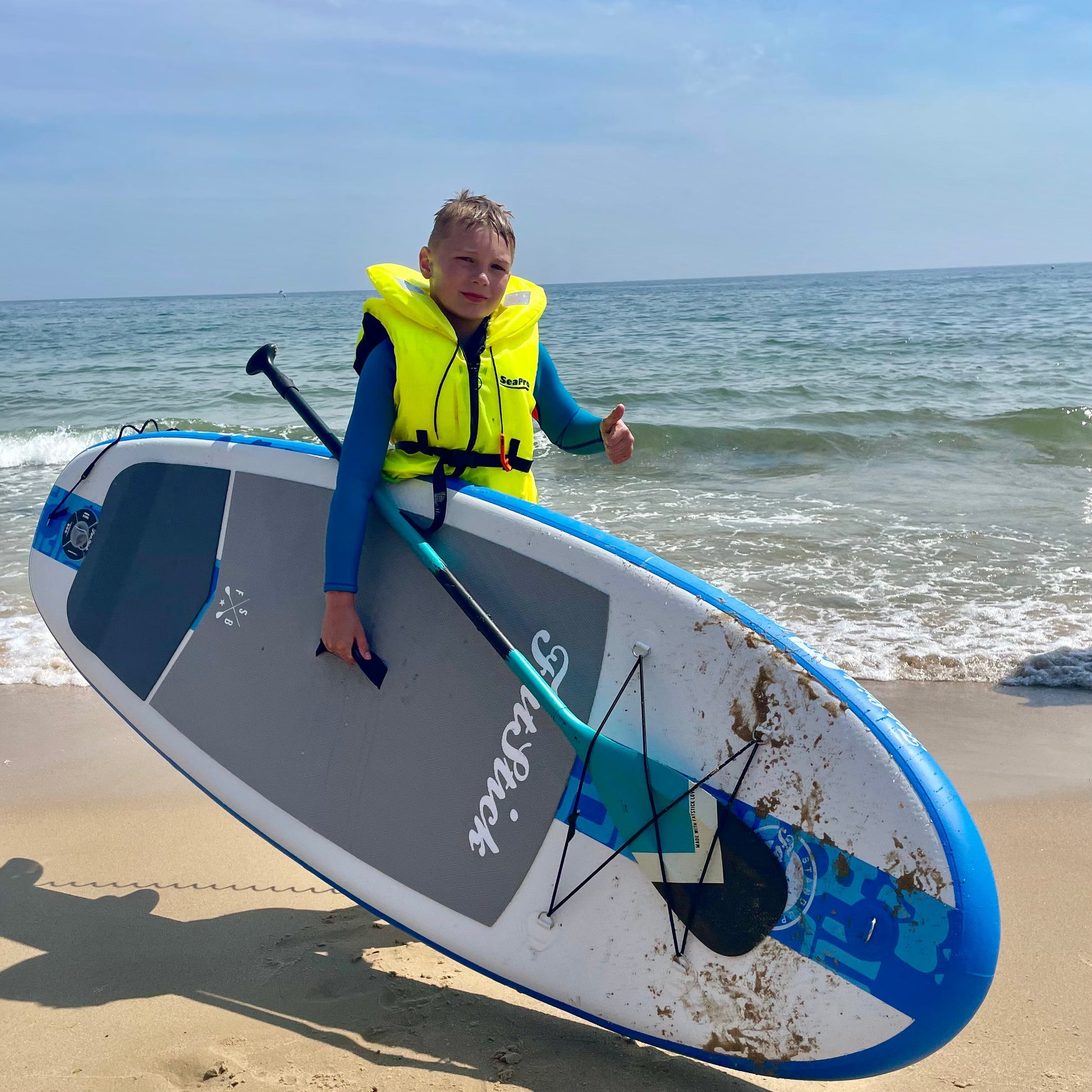 Young boy in a yellow life vest holds a blue Fatstick Gromstick inflatable paddle board on a sandy beach.