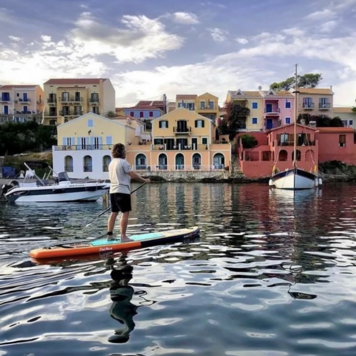 Man paddle boarding on the FatStick Wooden Menace 11'2 board in a calm bay with colorful buildings and boats in the background.
