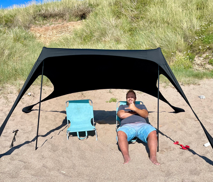 Man and blue beach chair under a black Fatstick Beach Tent sun shade on a sandy beach.