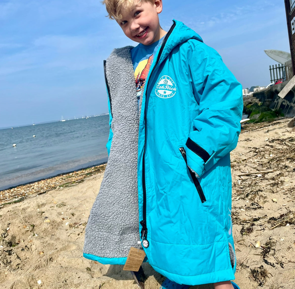 Young boy on beach wearing blue FatStick Kids FatWrap Changing Robe with full zipper open.