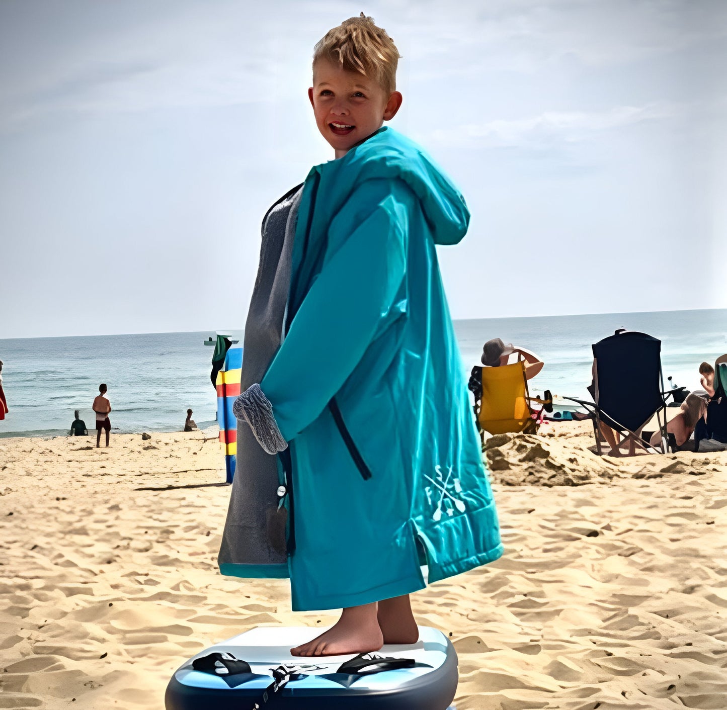Young boy in blue FatStick Kids FatWrap changing robe with sherpa fleece lining, smiling, on beach.