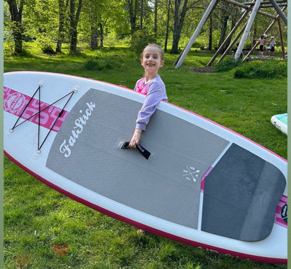 Smiling girl stands behind a pink Fatstick Gromstick inflatable paddle board with grass and trees behind.
