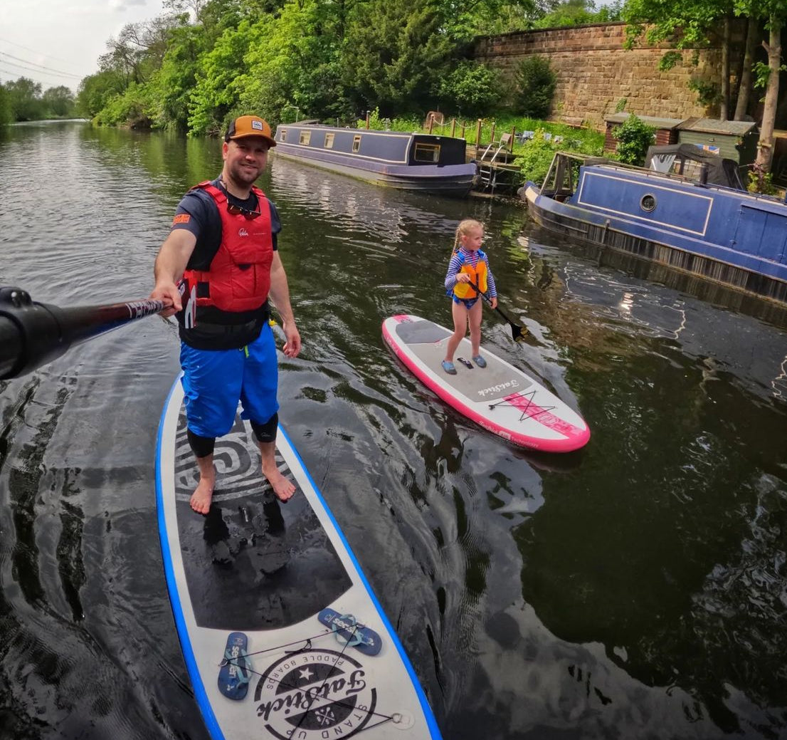 Adult and child on Fatstick inflatable paddle boards on a river with boats and trees.