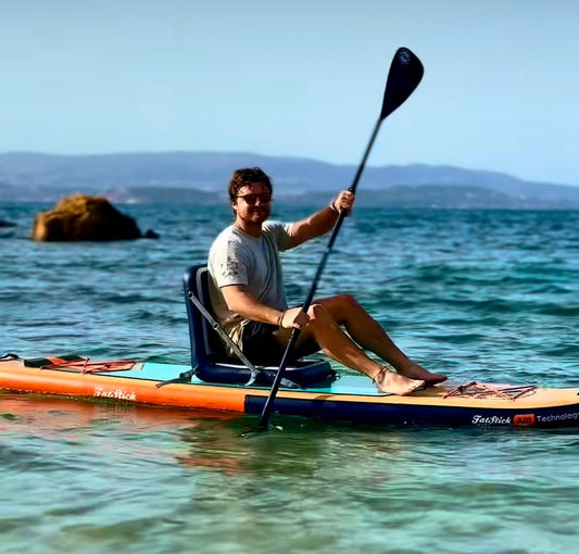 Man sitting on an orange paddle board with a black adaptive inflatable paddle board SUP kayak seat, paddling in clear blue water.