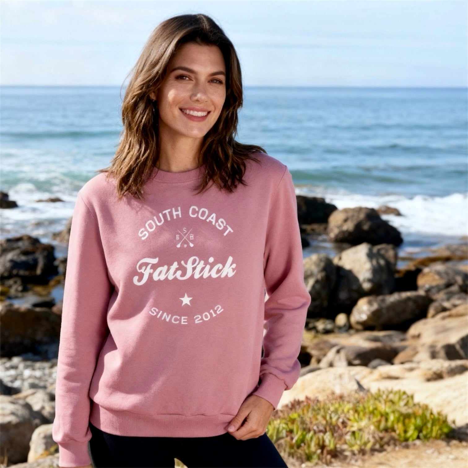 Woman in pink FatStick South Coast Lifestyle Sweater and dark pants smiling on a rocky beach.