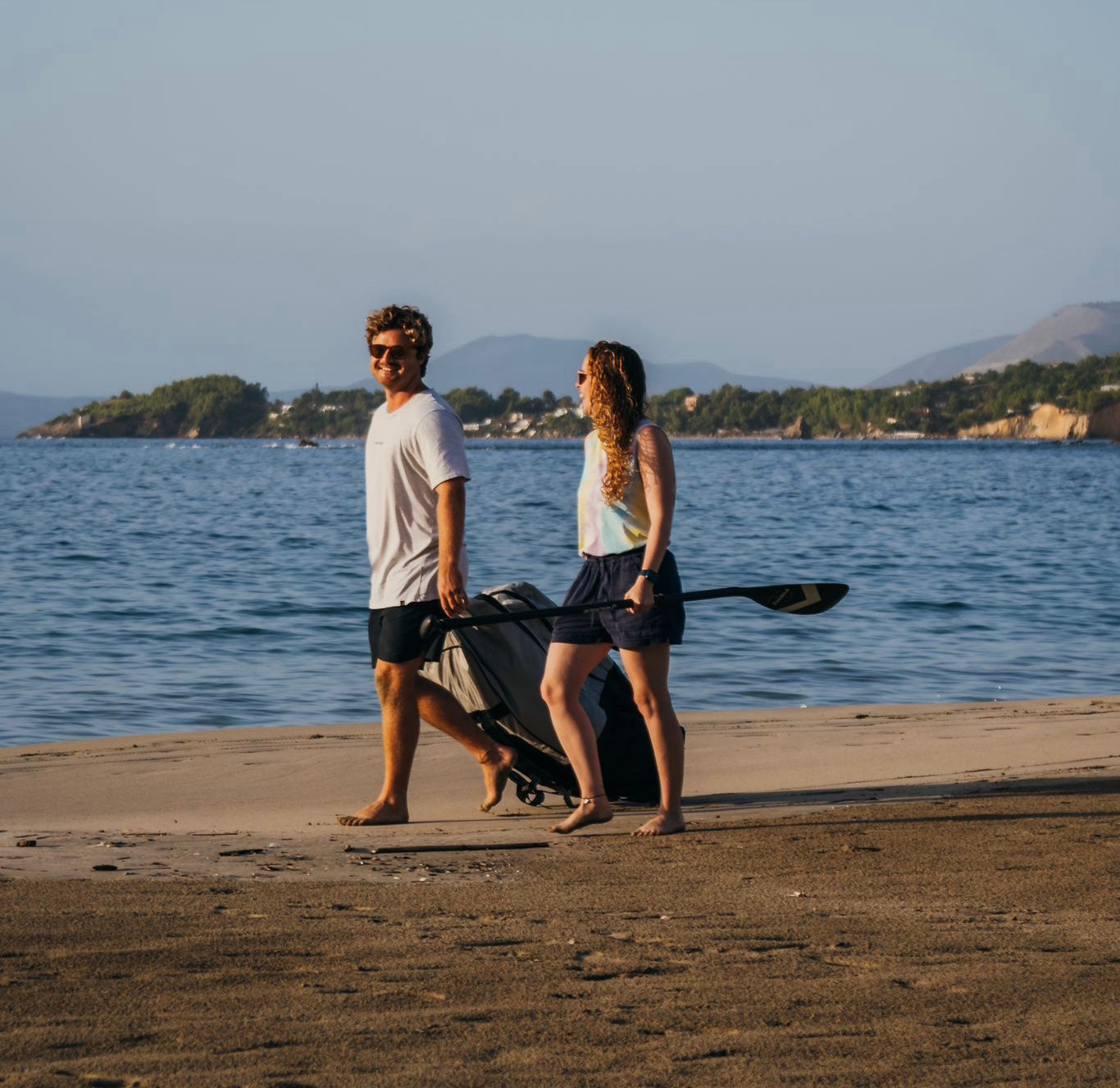 Man and woman walking on a beach, man pulling the FatStick Wheeled Storage Bag, woman holding a paddle.