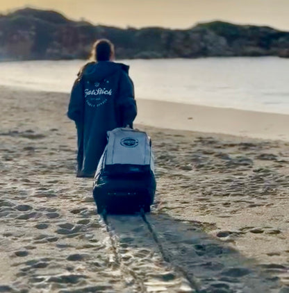 Woman in grey robe pulling a FatStick Wheeled Storage Bag Backpack on wheels along a sandy beach.