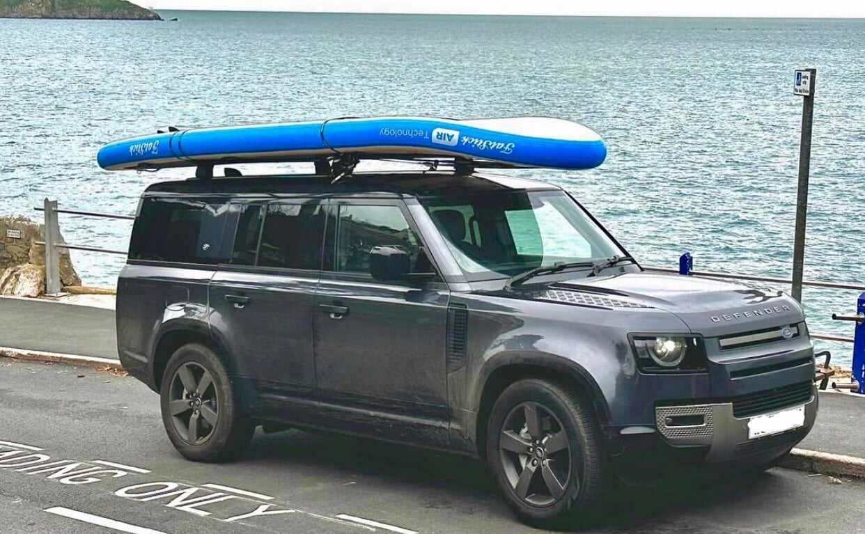 Side profile of an inflatable blue and white XL Giant FatStick Paddle Board Package strapped to the roof of a parked dark SUV.