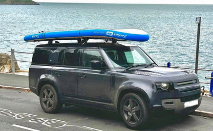 Side profile of an inflatable blue and white XL Giant FatStick Paddle Board Package strapped to the roof of a parked dark SUV.