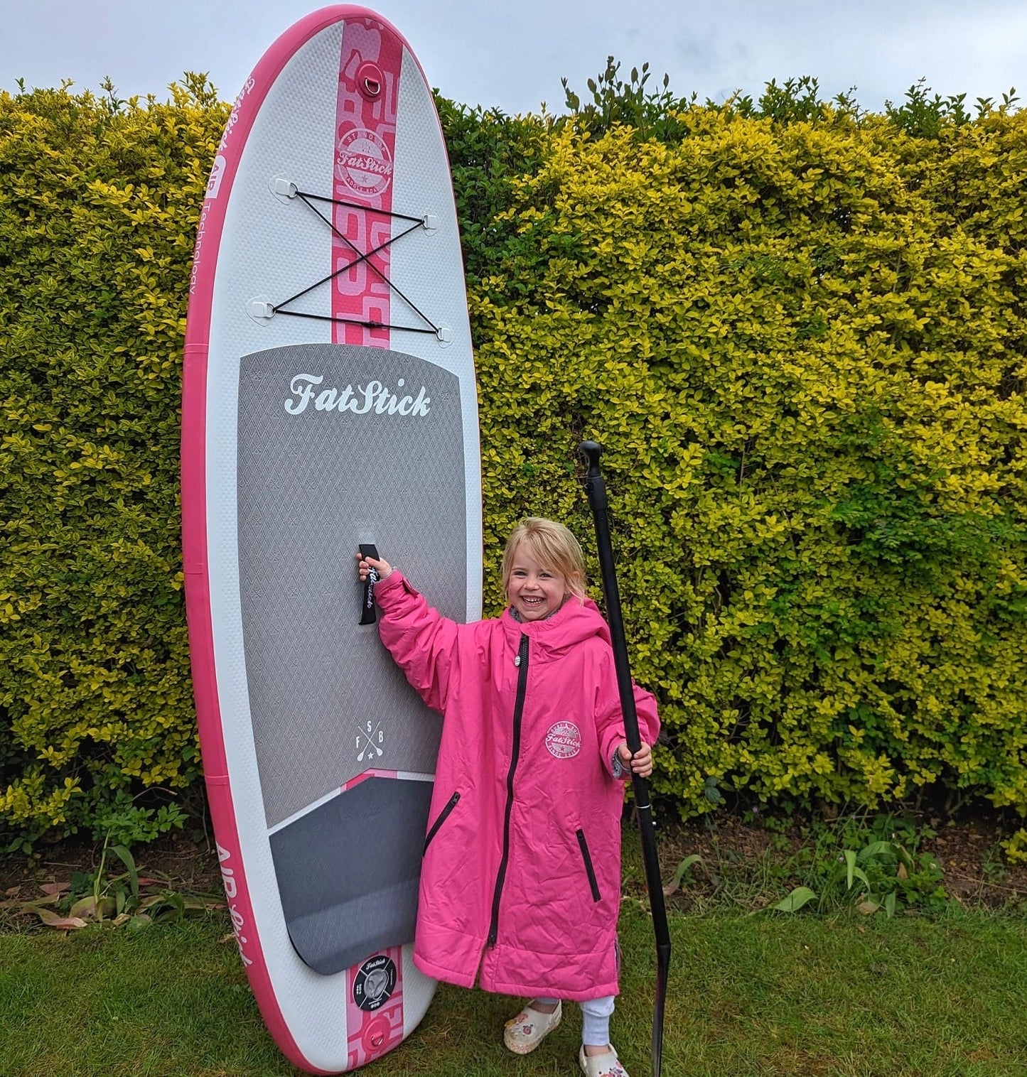 Smiling child in pink coat stands next to a pink Fatstick GromStick paddle board.