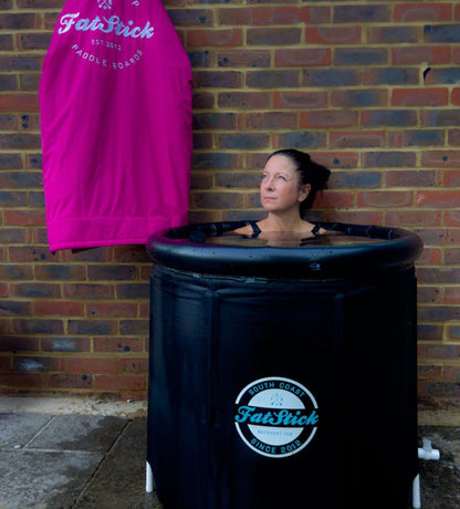 Woman shoulders-deep in a black FatStick Recovery Ice Bath XL outdoors against a brick wall.