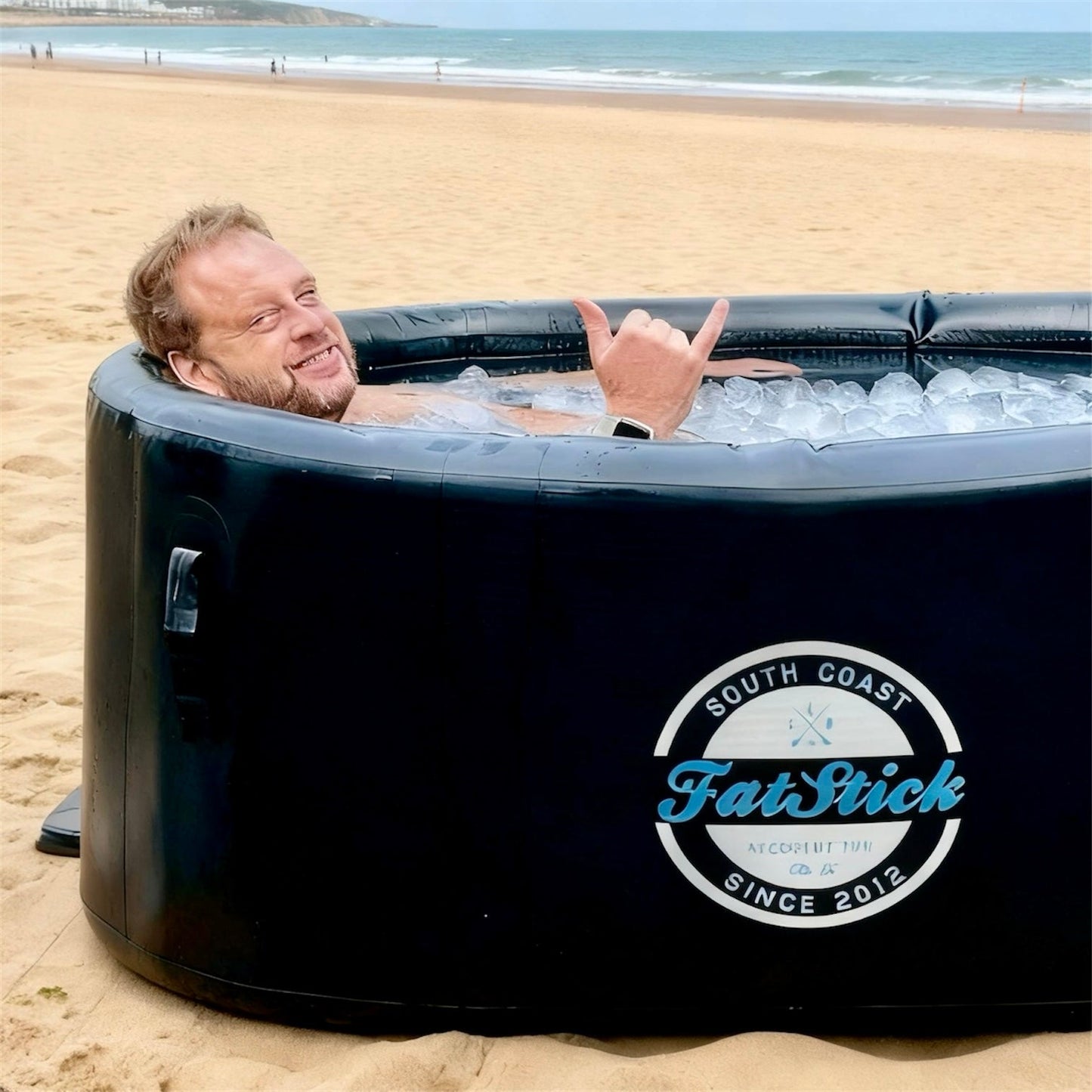Man in cold water therapy plunge pool on beach, ice bath filled with ice cubes, making shaka sign.