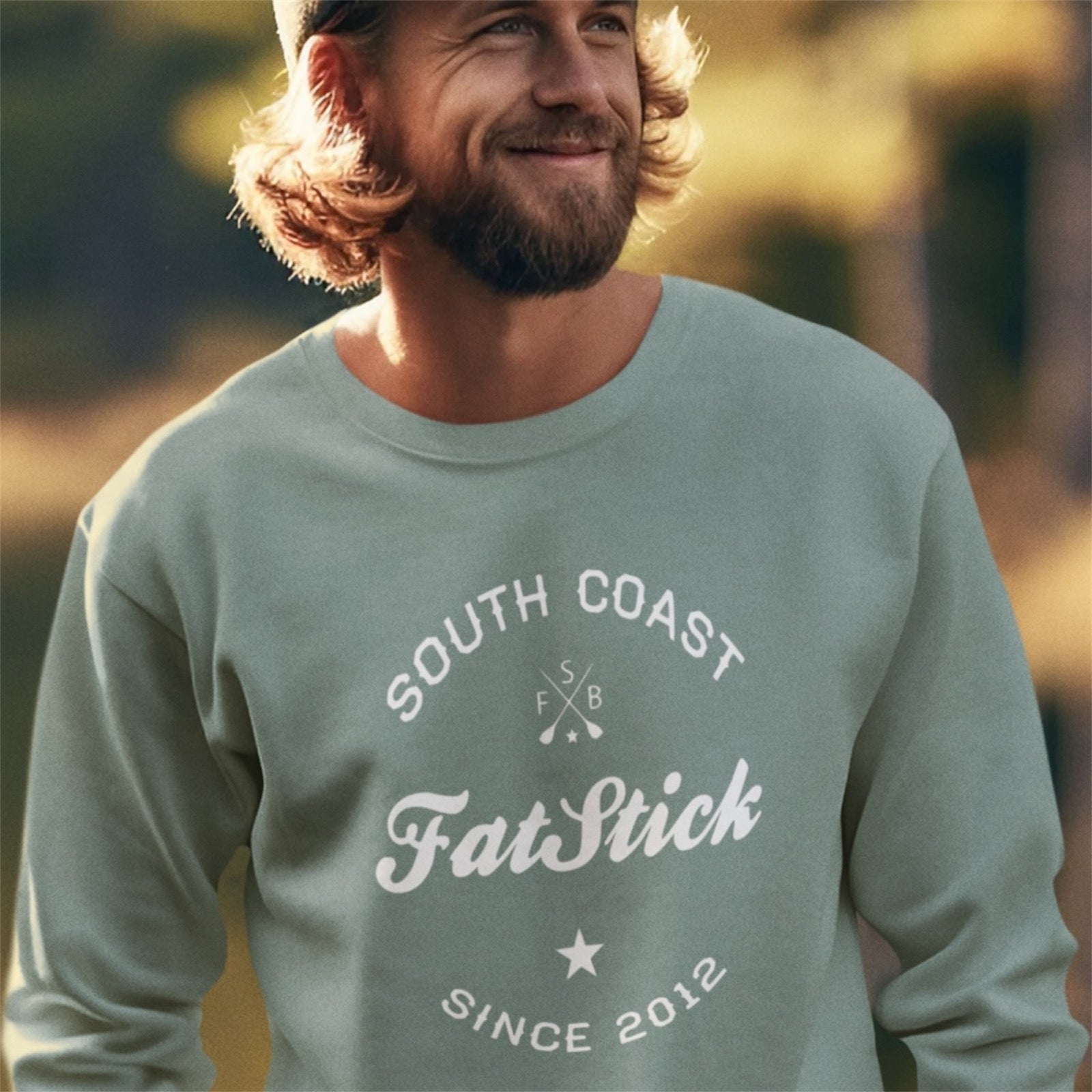 Smiling man wearing a light green FatStick South Coast Lifestyle Sweater outside in golden hour sun.