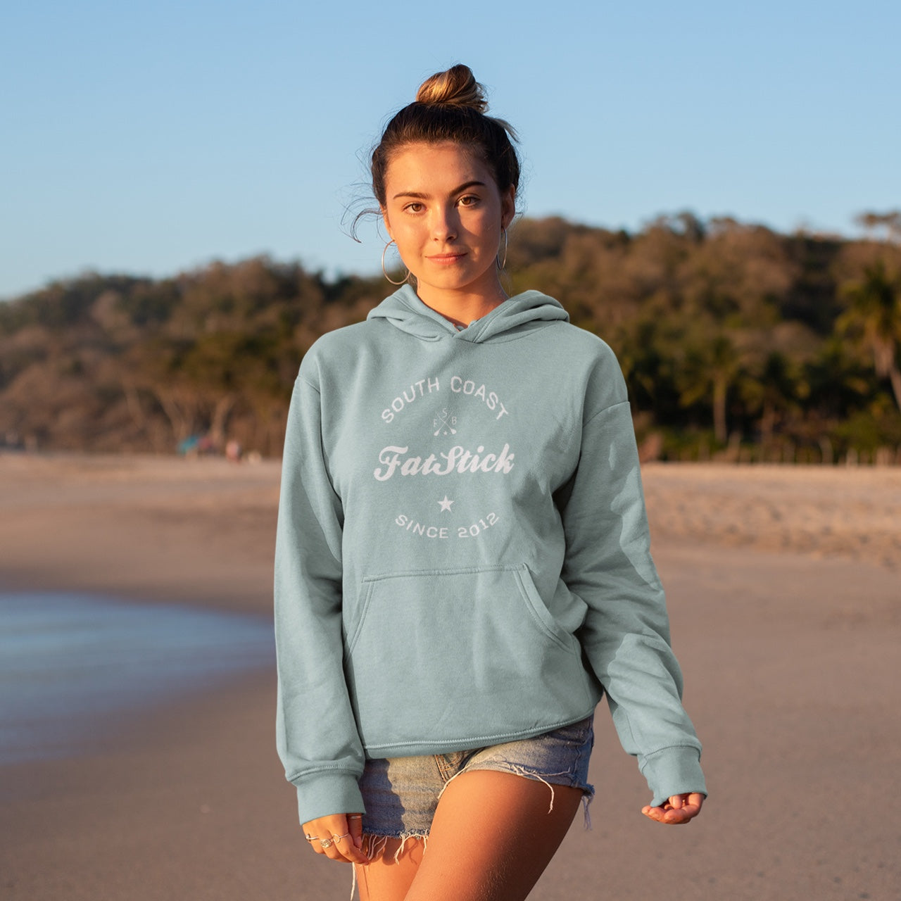 Young woman wearing a light blue FatStick South Coast Lifestyle Hoodie, standing on a beach with the ocean and green hills behind her