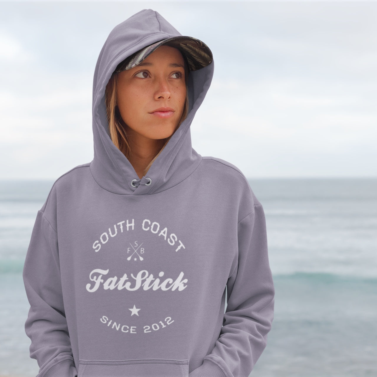 Young woman wearing a grey FatStick South Coast Lifestyle Hoodie with the hood up, looking right, with a blurred ocean background