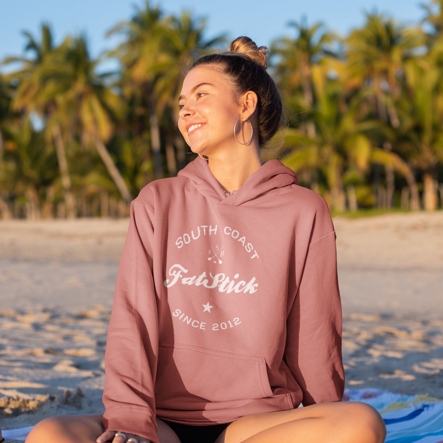 Young woman in a pink FatStick South Coast Lifestyle Hoodie sits on a beach facing right, smiling, with palm trees and ocean in the background