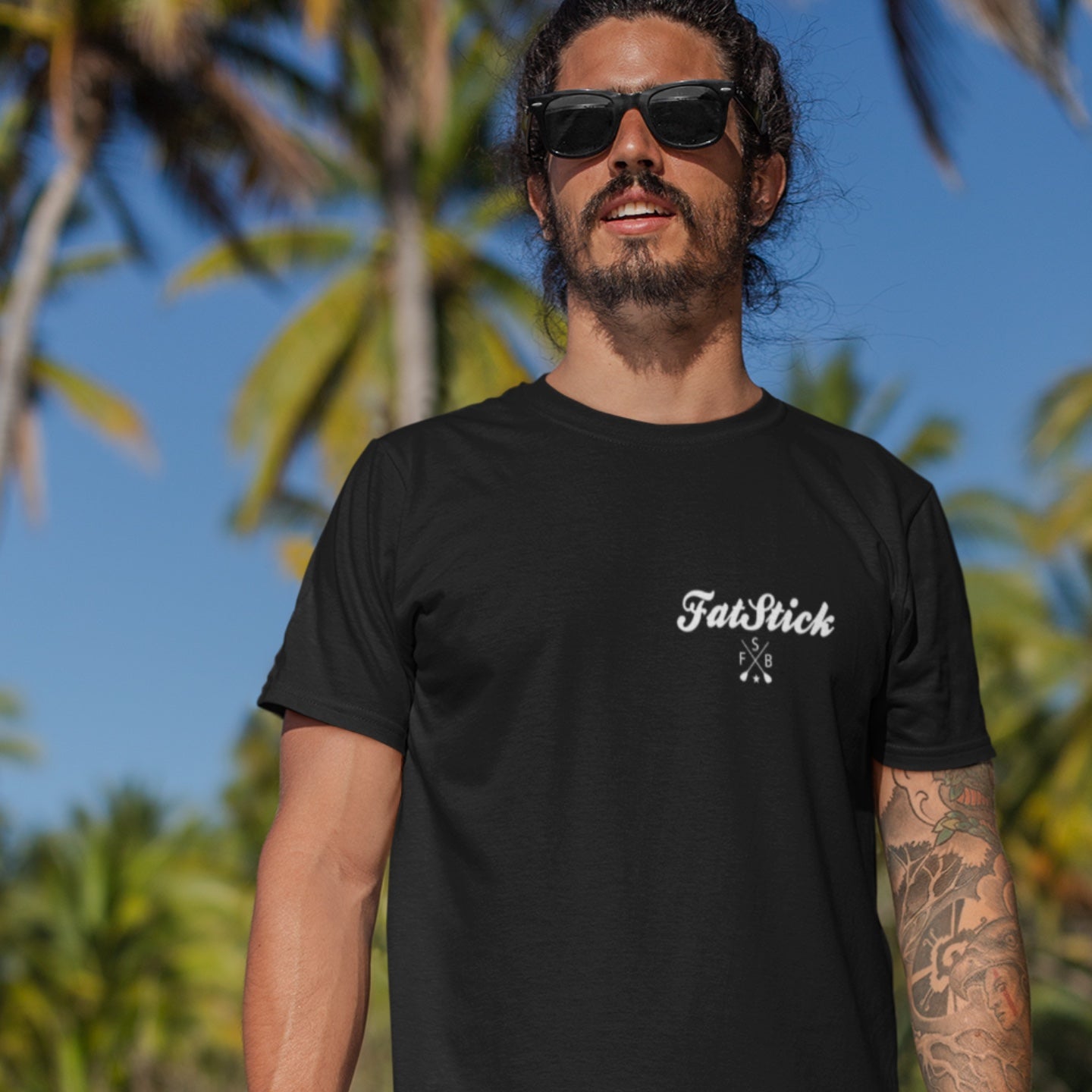 Man smiling wearing black FatStick South Coast Lifestyle T-Shirt and sunglasses, palm trees behind.