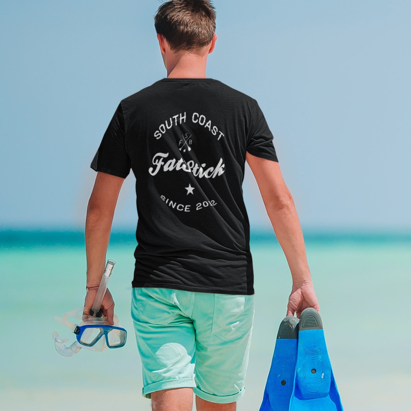 Man walking away on beach wearing black FatStick South Coast Lifestyle T-Shirt, holding snorkel gear.