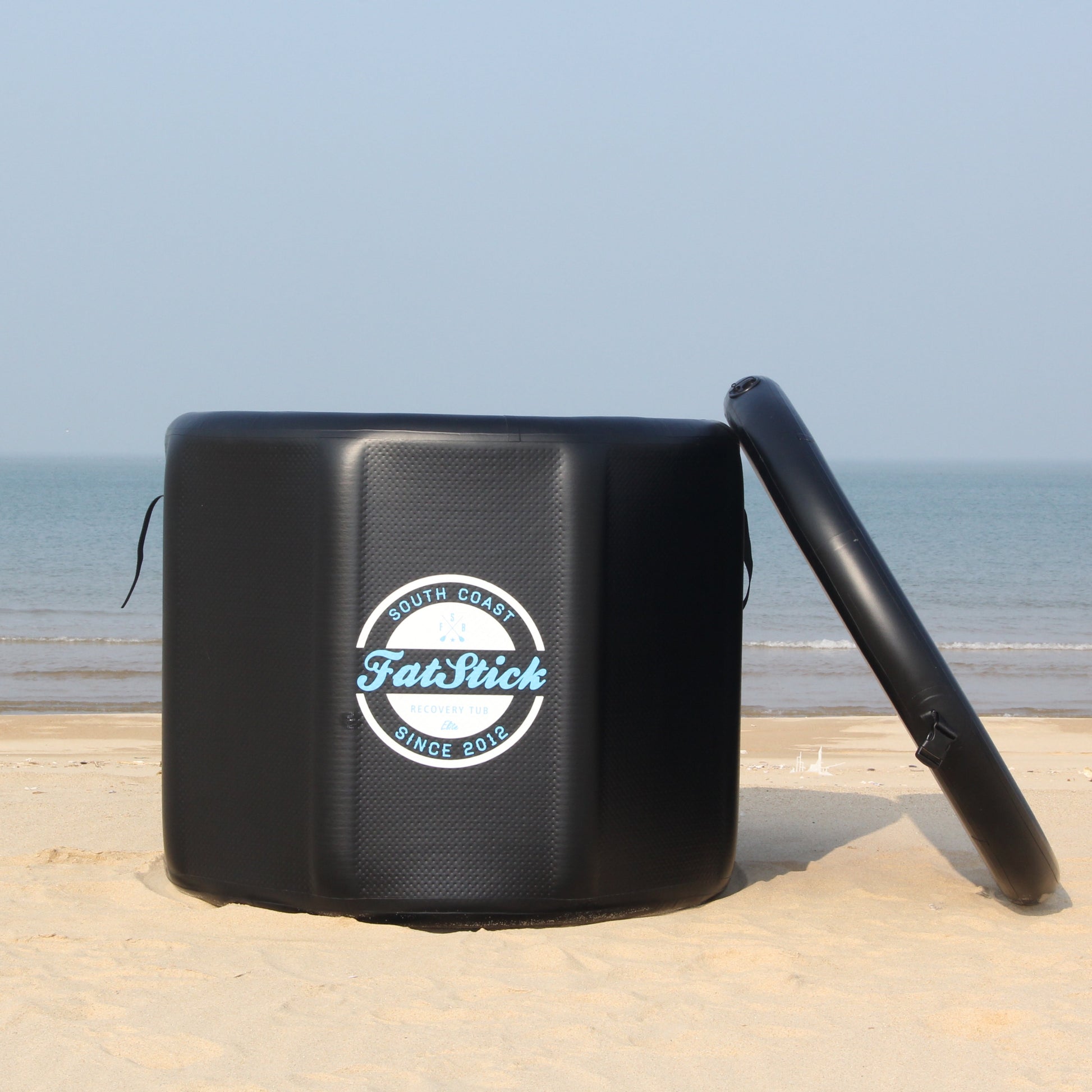 Round FatStick pro cold water therapy plunge tub on sand, with lid off to side, ocean background.
