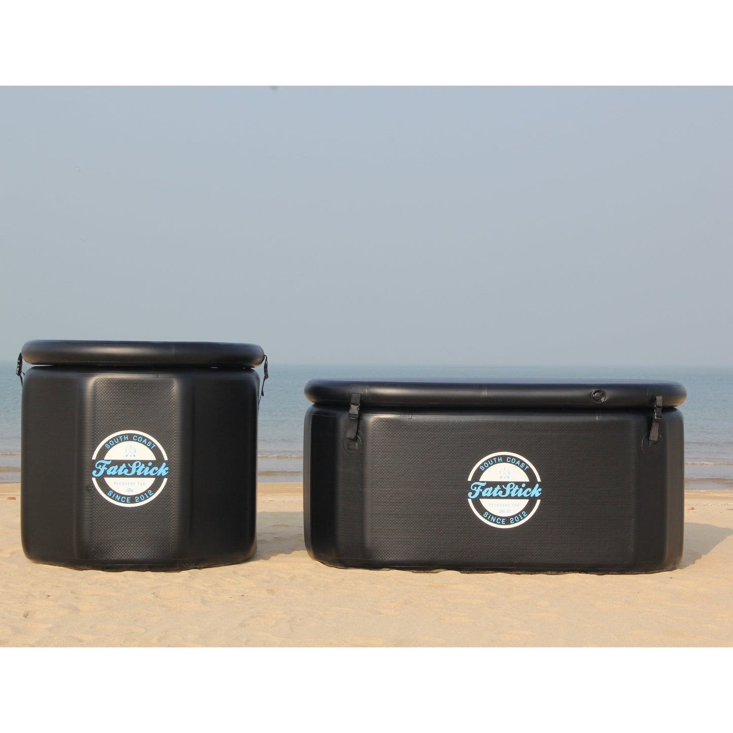 Two FatStick pro cold water therapy plunge tubs on the sand, an XL and standard size, ocean backdrop.