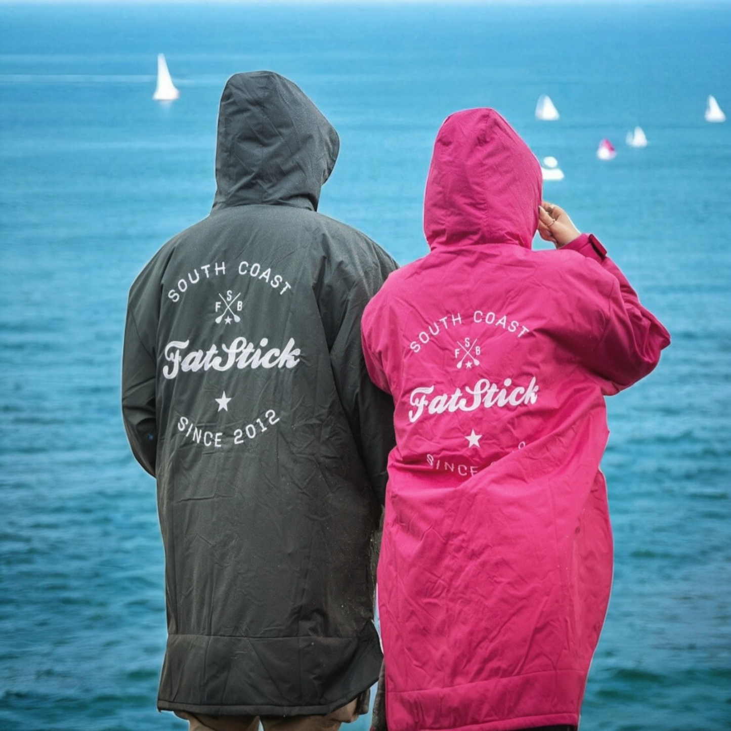 Rear view of two people in grey and pink FatWrap recycled fleece changing robes looking at the ocean and sailboats.