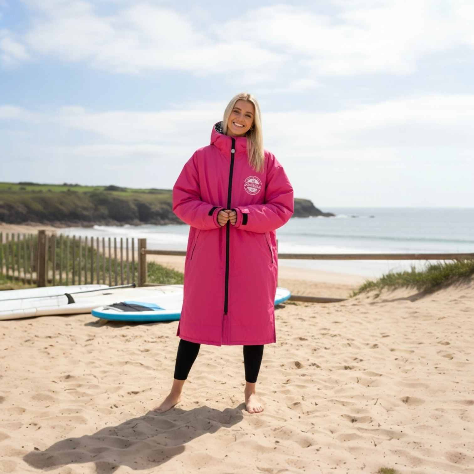 Front view of a smiling woman in a pink FatWrap recycled fleece changing robe standing on a sandy beach.