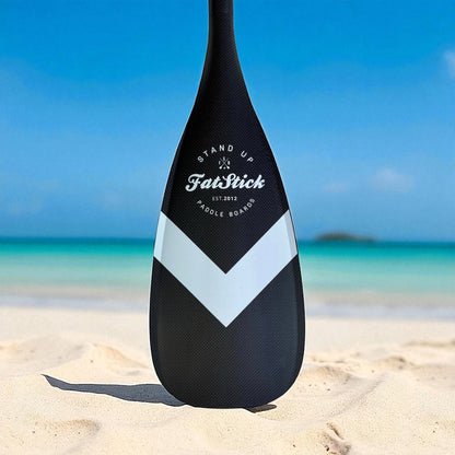 Close-up of FatStick full carbon paddle board blade, black with white chevron, on a sandy beach.