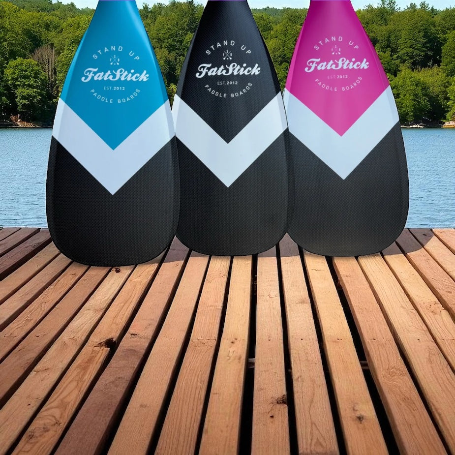 Three full carbon paddle board paddles in blue, black, and pink with white chevron design on a wooden dock overlooking a lake.