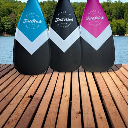 Three full carbon paddle board paddles in blue, black, and pink with white chevron design on a wooden dock overlooking a lake.