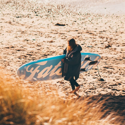 Woman carrying a blue AirStick 10'6 inflatable paddle board on a sandy beach with seagrass in the foreground.