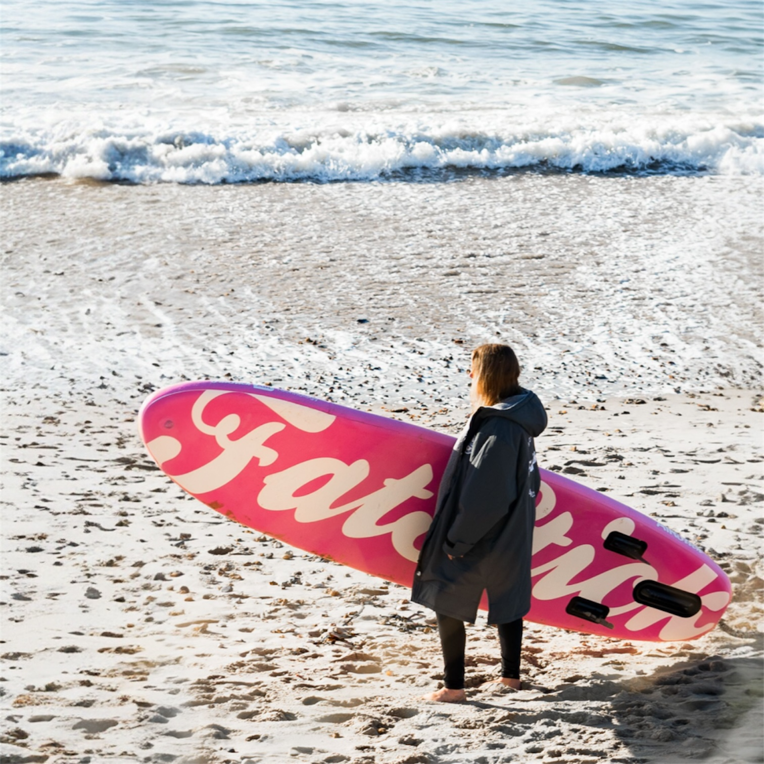 Rear view of a woman carrying a pink AirStick 10'6 inflatable paddle board on a sandy beach next to the ocean.