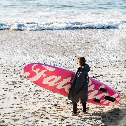 Rear view of a woman carrying a pink AirStick 10'6 inflatable paddle board on a sandy beach next to the ocean.
