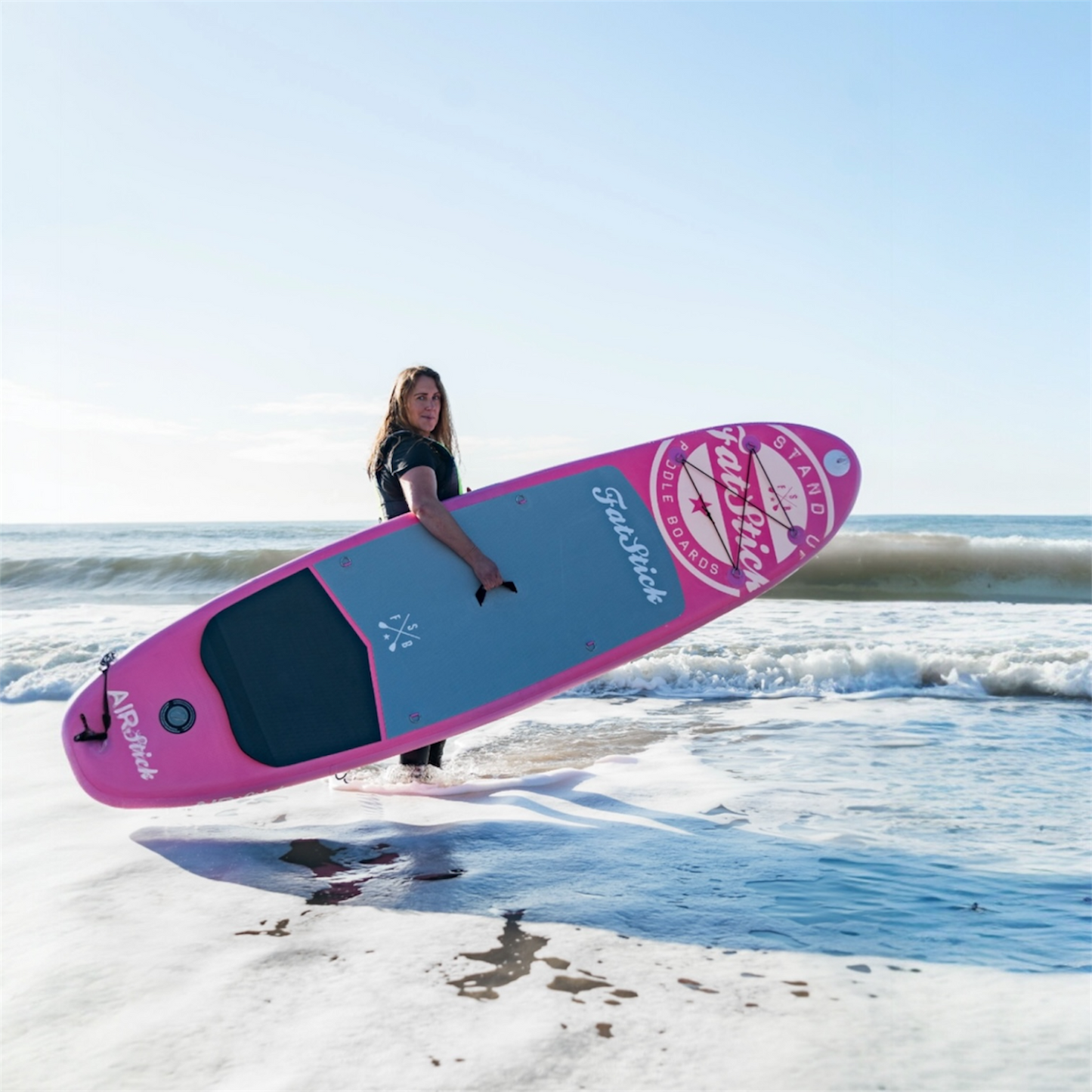 Woman in black wetsuit carrying a pink AirStick 10'6 inflatable paddle board through the shallow ocean water.