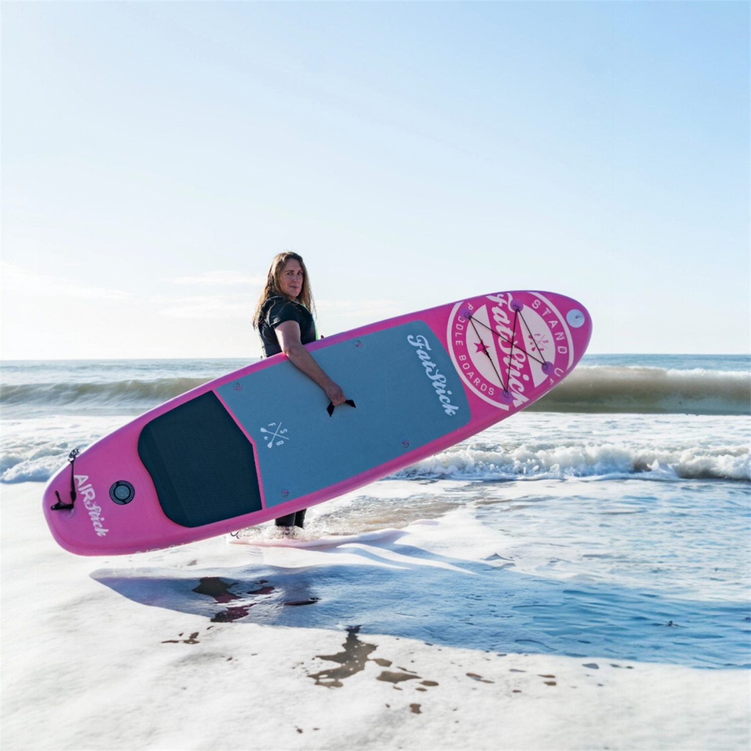 Woman in black wetsuit carrying a pink AirStick 10'6 inflatable paddle board through the shallow ocean water.