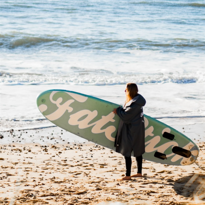 Woman in black wetsuit carrying a mint green AirStick 10'6 inflatable paddle board on a sandy beach.