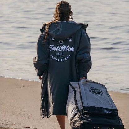 Rear view of model wearing a grey FatWrap recycled fleece changing robe, walking towards the ocean with a paddleboard bag.