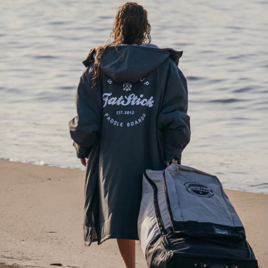 Rear view of model wearing a grey FatWrap recycled fleece changing robe, walking towards the ocean with a paddleboard bag.