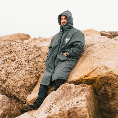 Front view of a man in a grey FatWrap recycled fleece changing robe sitting on large ocean rocks.