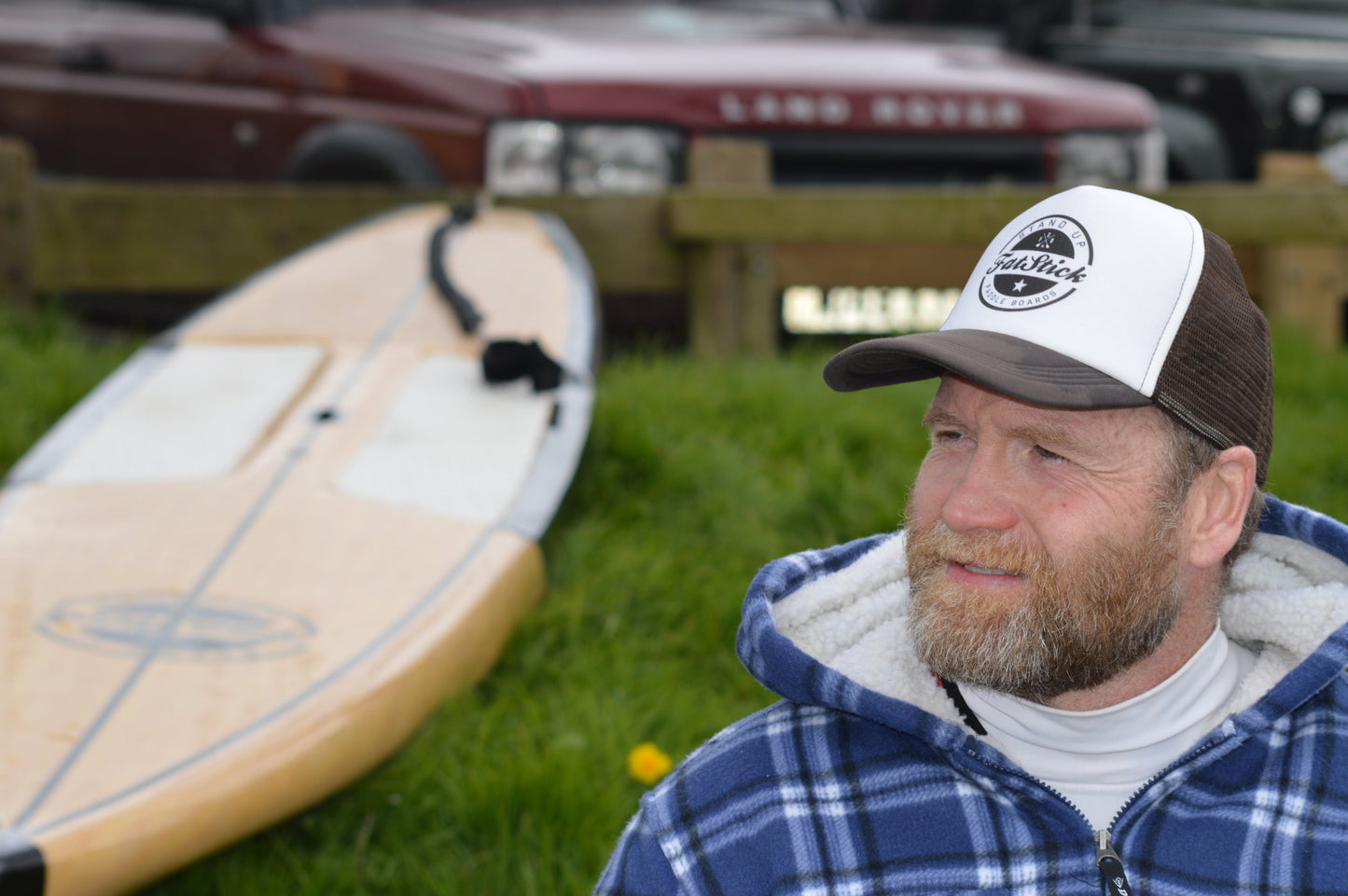 Man with a beard wearing a brown and white FatStick Team Trucker Hat next to a paddleboard
