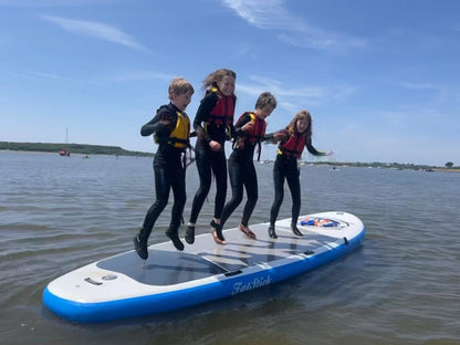 Four kids in wetsuits jumping simultaneously on the XL Giant FatStick Paddle Board package in open water under a clear sky.