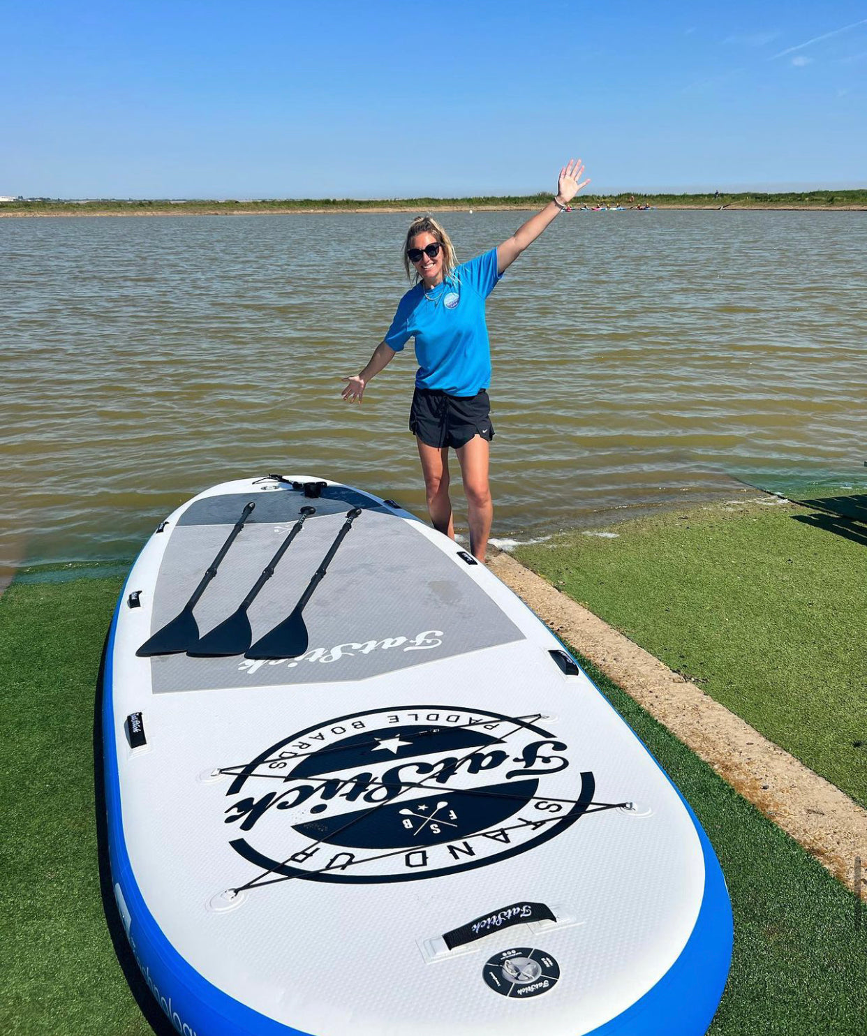 A woman in a blue shirt standing beside the XL Giant FatStick Paddle Board Package on green grass next to a lake, with paddles resting on the board.